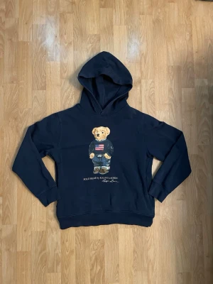 Marinblå hoodie Polo Bear Ralph Lauren - Snygg marinblå hoodie från Ralph Lauren med den ikoniska Polo Bear-trycket på bröstet. Hoodien har huva och ribbade muddar vid ärmar och nederkant. Perfekt för en avslappnad och trendig look. Materialet känns mjukt och bekvämt. Storlek 14-16 år barn. Sitter som XS