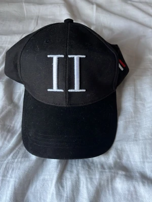 Svart keps från Tommy Hilfiger - Snygg svart keps från Tommy Hilfiger med stor vit broderad II-logga framtill och en liten röd, vit och blå detalj på sidan. Klassisk böjd skärm och justerbar baksida. Perfekt accessoar för en sportig och trendig look.