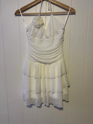 Vit kort volangklänning med en ros - Söt vit kort klänning med tunna axelband och halterneck. Klänningen har flera lager volanger i kjolen och en dekorativ blomrosett på bröstet. Draperad överdel och lätt, flowigt tyg som ger en romantisk känsla. Använts en gång. Pris kan diskuteras!