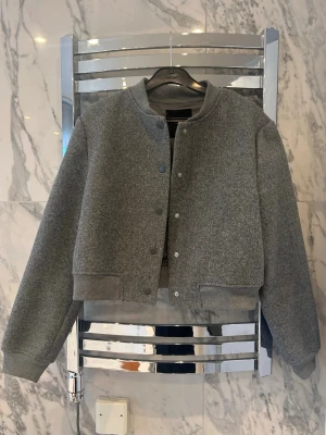 Grå cropped bomberjacka - Stilren grå bomberjacka i cropped modell med rund halsringning och tryckknappar framtill. Jackan har breda muddar vid ärmslut och nederkant för en sportig look. Tillverkad i ett mjukt, tjockare tyg som ger en avslappnad vibe.