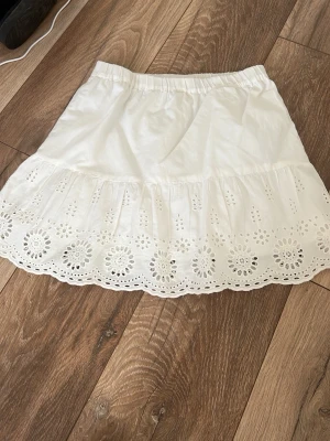 Vit kortkjol med broderad volang Kappahl - Söt vit kortkjol från Kappahl med bred volangkant och broderade blommönster. Kjolen har resår i midjan och är gjord i ett lätt bomullstyg, perfekt för varma dagar. Passar dig som gillar romantisk och somrig stil.