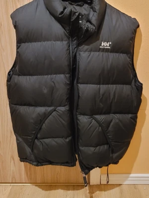 Svart dunväst från Helly Hansen - Snygg svart dunväst från Helly Hansen med hög krage och dragkedja framtill. Västen har två stora fickor med dragkedja och logga på bröstet. Perfekt för lager-på-lager och håller dig varm under kyliga dagar. Klassisk puffer-look och justerbar nederkant.