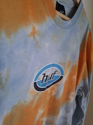 HUF tie-dye långärmad topp - Snygg långärmad topp från HUF med tie-dye mönster i orange och ljusblått. Stor HUF-logga framtill. Perfekt för dig som gillar streetwear och vill sticka ut med färg. Mjuk och skön bomullskvalitet.