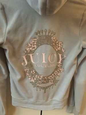 Ljusblå Juicy Couture zip - Snygg ljusblå hoodie från Juicy Couture i mjuk velour. Dragkedja framtill, två fickor och huva med snören. Stor logga i silver och rosa med krona och blommor på ryggen. Perfekt för en chill och trendig look.