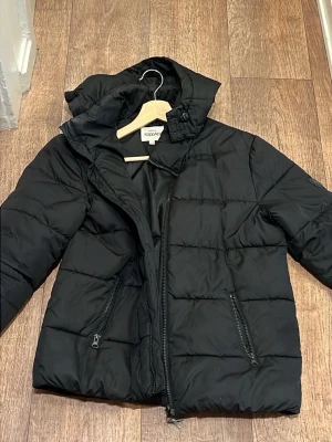 Svart pufferjacka från Ocean District - Svart pufferjacka från Ocean District med huva och två dragkedjeförsedda fickor. Jackan har quiltad design och är perfekt för kalla vinterdagar. Materialet är syntet och jackan har en stilren look med en diskret logotyp på ärmen.