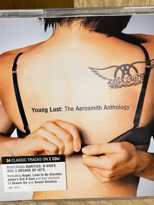 Aerosmith Young Lust Anthology CD - Young Lust: The Aerosmith Anthology är en dubbel-CD med 34 klassiska låtar från Aerosmith. Omslaget har en svartvit bild med Aerosmiths logga och låtlista på baksidan. Perfekt för dig som gillar rock och samlar på musik.