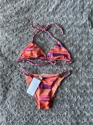 Färgglad bikini från Mango - Snygg bikini som är slutsåld från Mango med färgglada vågiga ränder i orange, rosa och lila toner. Bikinin har trekantsöverdel med knyt i nacken och baktill samt matchande underdel med flätade band i sidorna. Har inlägg i överdelen (limited edition!!)🧡🩷💜