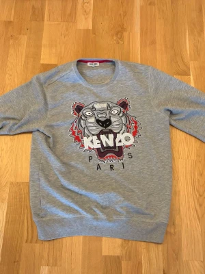 Grå sweatshirt från Kenzo med tiger - Säljer en grå sweatshirt från Kenzo med det ikoniska tigermotivet broderat framtill och texten 'KENZO PARIS'. Tröjan har rund halsringning, ribbade muddar och är tillverkad i mjuk bomullsblandning. Perfekt för dig som gillar streetwear och vill sticka ut.