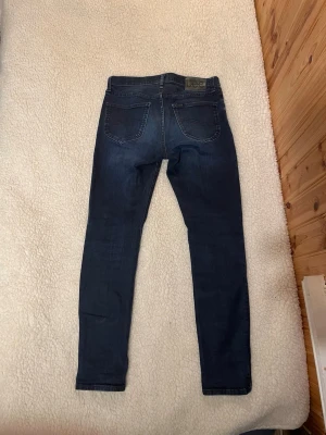 Mörkblå  jeans från Lee - Snygga mörkblå jeans från Lee med klassisk femficksdesign och diskreta slitningar. Jeansen har normal midja och smal passform hela vägen ner. Tillverkade i stretchigt denim för extra komfort och rörelsefrihet. W29 och L32.