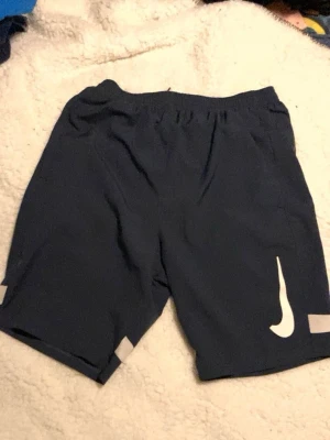 Blå Nike träningsshorts - Mörkblå träningsshorts från Nike med elastisk midja och vita detaljer, inklusive den klassiska Nike-loggan på ena benet och vita ränder längst ner. Perfekta för sport och aktivitet.