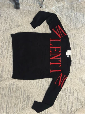 Svart stickad tröja från Valentino - Snygg svart stickad tröja från Valentino med stora röda bokstäver över bröstet och ärmarna. Tröjan har rund halsringning och långa ärmar. Perfekt för dig som gillar statement-plagg och exklusiv design.