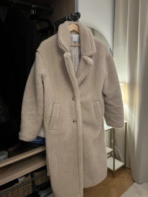 Beige lång teddyjacka med krage - Säljer en beige teddyjacka i lång modell med bred krage och två framfickor. Jackan har knäppning framtill och är gjord i mjukt, fluffigt material som ger en riktigt cozy vibe. Perfekt för kalla dagar när du vill vara både varm och stylish. Passar xs - s