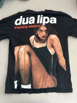 Svart Dua Lipa t-shirt Training Season - Cool svart t-shirt med stort tryck av Dua Lipa och texten 'training season' i vitt och rött. Klassisk passform med korta ärmar och rund hals. Perfekt för dig som gillar musik och vill ha en unik merch-tröja.