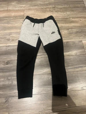 Nike Tech Fleece mjukisbyxor svart/grå - Nike Tech Fleece mjukisbyxor i svart och ljusgrått med dragsko i midjan och ribbade muddar vid benslut. Byxorna har fickor fram och en diskret Nike-logga på vänster lår. Perfekta för en avslappnad och sportig stil.