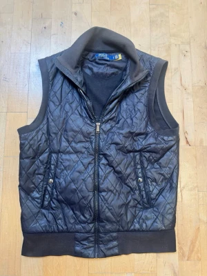 Ralph Lauren väst  - Snygg svart dunväst från Polo Ralph Lauren med quiltat mönster framtill, mjuk krage och ribbade detaljer vid ärmhål och nederkant. Två fickor med tryckknappar och dragkedja framtill. Perfekt för våren och sommarkvällar. Gått upp i sömmarna innanför dragkedjan (se sista bilden).  hör av dig vid minsta funderingar eller frågor!😊