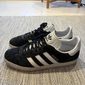 Adidas Gazelle svartvita sneakers - Klassiska Adidas Gazelle sneakers i svart mocka med vita detaljer och de ikoniska tre ränderna på sidorna. Skorna har vit plös och häl med Adidas-logga, samt svart snörning och platt sula. Perfekta för en stilren och sportig look.