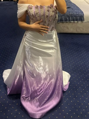 Långklänning i lila och vitt med spets - Magisk långklänning med offshoulder och vacker ombre-effekt i vitt och lila. Livet är dekorerat med spets och pärlor samt har en romantisk snörning. Kjolen är glansig och svepande, perfekt för speciella tillfällen. Armarna kan man ta bort 