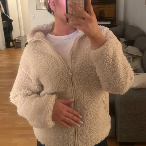 Krämvit teddyjacka med dragkedja - Mysig teddyjacka i krämvit färg med hög krage och hel dragkedja framtill. Jackan har en fluffig och mjuk struktur, perfekt för att hålla värmen. Passar dig som gillar en avslappnad och trendig stil. Materialet är troligtvis polyester.