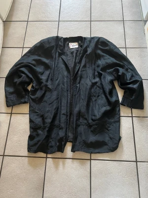 Svart oversized cardiganjacka i siden - Snygg svart oversized cardiganjacka i tunt, glansigt sidenmaterial. Jackan har avslappnad passform, vida ärmar och öppen front utan knappar. Perfekt för en chill och stilren look. Se bilder för skick 