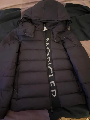 Kom me prisförslag!!! Svart dunjacka från Moncler - Säljer en svart dunjacka från Moncler med stor huva och tydlig logotyp längs dragkedjan. Jackan har quiltad design och är tillverkad i glansigt material med certifierat dun. Perfekt för kalla vinterdagar.