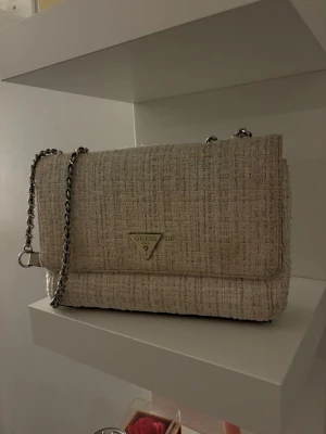 Beige axelväska från Guess i tweed - Snygg beige axelväska från Guess i tweedmaterial med silverfärgad kedjerem och klassisk trekantslogga framtill. Väskan har en stilren, rektangulär form och detaljer i ljusbeige konstläder. Perfekt för dig som gillar trendiga accessoarer.