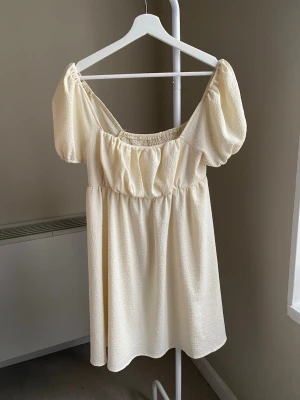 Ljus kort klänning med puffärm - Supersöt ljus kort klänning med puffärmar och smockad överdel. Klänningen har en luftig passform och är tillverkad i ett strukturerat tyg som känns mjukt mot huden. Perfekt för varma dagar och ger en romantisk vibe.