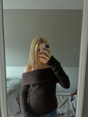 Brun off-shoulder stickad tröja - En mörkbrun stickad tröja med off-shoulder design och långa ärmar. Tröjan har en bred krage som går över axlarna och är ribbstickad för en snygg och trendig look. 