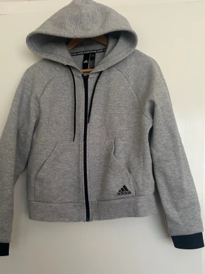 Grått Adidas mjukisset - Säljer ett grått mjukisset från Adidas i väldigt bra skick då den tyvärr inte kommer till användning.  Hoodie med dragkedja och matchande mjukisbyxor med de klassiska tre ränderna.  Kofta i S. Byxorna är märkta 13–14 years men motsvarar ungefär XS/S.
