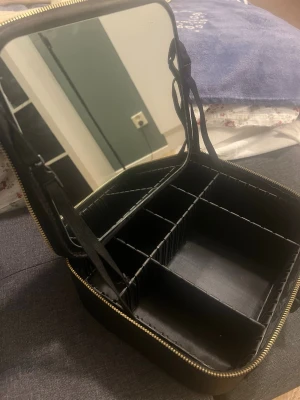 Svart sminkväska med spegel - Praktisk svart sminkväska med flera fack och justerbara avdelare. Väskan har en stor spegel i locket och elastiska fickor för småsaker. Perfekt för att organisera smink eller accessoarer. Stängs med dragkedja och har en stilren design.