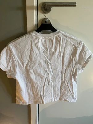 Vit croppad t-shirt från Lyle & Scott - Säljer en vit croppad t-shirt från Lyle & Scott med klassisk gul logga på bröstet. T-shirten har korta ärmar, v-ringning och är gjord i mjuk bomull. Perfekt till jeans eller shorts för en clean och enkel look.