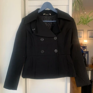 Svart dubbelknäppt skepparkavaj, Tiger of Sweden - Stilren svart kappa/ skepparkavaj med dubbelknäppning och bred krage. Kortare modell med markerad midja och två fickor framtill. Fodrad insida för extra komfort. Perfekt för dig som vill ha en klassisk och tidlös look.