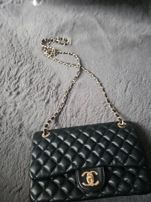 Svart quiltad handväska - Säljer en klassisk svart quiltad handväska från med kedjerem i guld och ikonisk CC-logo på framsidan. Väskan är tillverkad i läder och har en elegant, tidlös design med lock och ficka på baksidan.