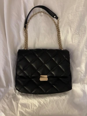 Svart quiltad handväska med guldkedja - Snygg svart handväska i quiltat mönster med guldfärgad kedjerem och läderdetalj upptill. Väskan har en rektangulär form och stängs med ett stilrent guldfärgat spänne framtill. Perfekt accessoar för att lyfta din outfit.