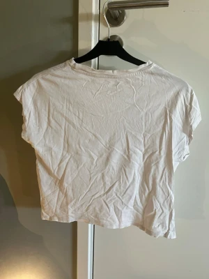 Vit croppad t-shirt från H&M - En enkel vit croppad t-shirt från H&M med korta ärmar och rund halsringning. Perfekt för en avslappnad stil och lätt att matcha med jeans eller kjol. Tillverkad i mjuk bomull för en bekväm känsla.