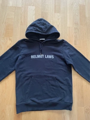 Svart hoodie Helmut Lang - Svart hoodie med stor huva och dragsko, samt magficka framtill. På bröstet står det 'HELMUT LAWS' i vitt tryck och på ryggen finns ytterligare vitt tryck med text. Tillverkad i mjukt bomullsmaterial, perfekt för en avslappnad streetwear-look.