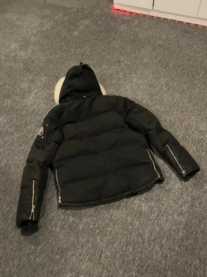 Svart pufferjacka från Moose Knuckles - Säljer en svart pufferjacka från Moose Knuckles med ikonisk logga i metall på ärmen. Jackan har huva med vit fuskpäls, dragkedjor vid ärmslut och sidor samt en cool, vadderad look. Perfekt för kalla vinterdagar och riktigt snygg streetstil.