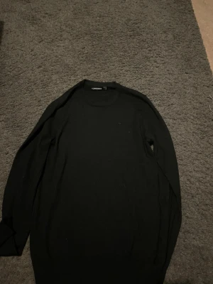 Svart stickad tröja från j.lindberg - En klassisk svart stickad tröja från J.lindeberg med rund halsringning och långa ärmar. Tröjan är enkel i modellen och passar perfekt till många olika stilar. Materialet ser ut att vara mjukt och bekvämt, perfekt för lager-på-lager.