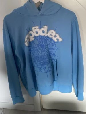 Sp5der hoodie - Säljer en blå hoodie från Sp5der med ett stort spindelnätsmotiv och broderad logga på bröstet. Hoodien har huva, ribbade muddar och är gjord i mjukt bomullsmaterial. Perfekt för dig som gillar streetwear och vill sticka ut med en unik design.