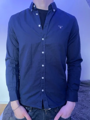 Mörkblå skjorta från Gant - Snygg mörkblå skjorta från Gant med button-down krage och vita knappar. Skjortan har lång ärm och diskret Gant-logga broderad på bröstet. Tillverkad i ett mjukt bomullstyg som känns bekvämt och stilrent.