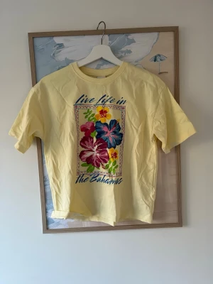 Gul Bahamas t-shirt med blommor - Gul t-shirt med färgglatt blommönster och texten 'live life in the Bahamas' på framsidan. T-shirten har korta ärmar och en klassisk passform. Perfekt för dig som gillar retrovibbar och somriga prints.