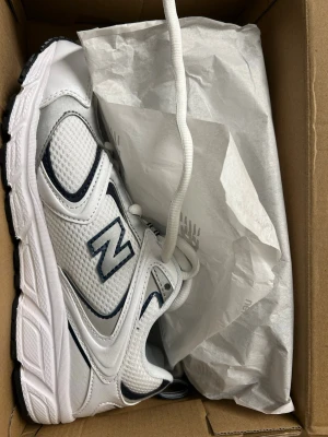 Vita New Balance sneakers - Snygga vita sneakers från New Balance med marinblå detaljer. Skorna har en klassisk låg siluett, ovandel i mesh och syntetmaterial samt tydlig N-logga på sidan. Perfekta för dig som gillar sportig och clean stil.