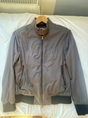 Grå bomberjacka från Oscar Jacobson - Snygg grå bomberjacka från Oscar Jacobson med svarta muddar och dragkedja framtill. Jackan har två fickor med dragkedja och en hög krage med tryckknapp. Perfa för våren 