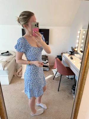 Blå blommig kortklänning med puffärm - Söt blå kortklänning med vita blommor och puffärmar. Klänningen har en v-ringad hals och figurnära passning som framhäver siluetten. Perfekt för sommarens dagar och har en lätt och luftig känsla. Materialet känns som bomull eller polyester.