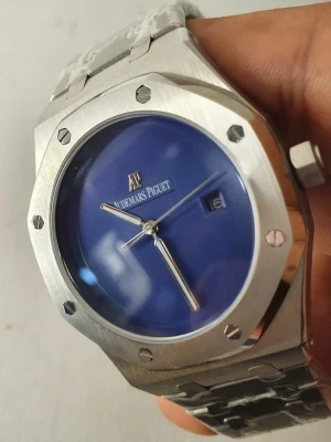 Audemars Piguet Royal Oak blå klocka - Snygg Audemars Piguet Royal Oak klocka med blå urtavla och datumvisning. Klockan har en ikonisk åttakantig boett i silverfärgat stål och matchande länkarmband. Minimalistisk design med tydliga visare och AP-logga på urtavlan. Perfekt för dig som gillar exklusiva accessoarer.