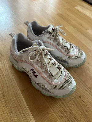  Fila sneakers - Snygga vita Fila sneakers med rosa och turkosa detaljer. Skorna har chunky sula, Fila-logga i vinrött på sidan. Perfekta för en sportig och trendig look.