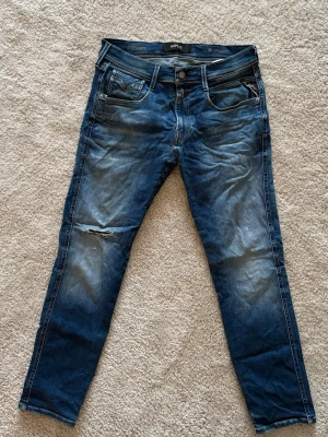 Replay Jeans - Säljer ett par blå jeans från Replay med skinny fit och slitningar på vänster lår. Jeansen har klassisk femficksmodell, dragkedjegylf och snygga tvättade detaljer. Perfekta för en avslappnad och trendig look.