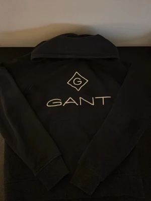 Marinblå hoodie från GANT med logga - Svart hoodie från GANT med stor vit broderad logga och emblem på bröstet. Klassisk modell med huva och känguruficka framtill. Mjuk bomullsblandning och ribbade muddar vid ärmslut och nederkant. Perfekt för en avslappnad och stilren look. Notera en fläck på magen, se sista bilden