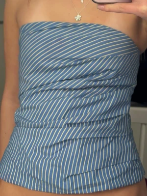 Blåvit randig tubtopp - Snygg blå tubtopp med vita tunna ränder. Toppen är axelbandslös och har en bandeauformad passform som sitter tight över bröstet. Perfekt för varma dagar och enkel att styla med jeans eller kjol.