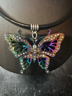 Regnbågsfärgat fjärilshalsband - Unikt halsband med en stor fjärilshängare i regnbågsfärgad metall och gnistrande strasstenar. Hängaren har detaljer i grönt, blått, lila och gult samt svarta inslag. Kedjan är i svart läderrem  och fjärilen är dekorerad med klara stenar för extra bling.