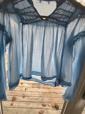 Ljusblå transparent blus med spetsdetaljer - Snygg ljusblå blus i transparent tyg med dekorativa spetsdetaljer vid axlar och ärmar. Blusen har långärmad design och ett luftigt, flowy fall. Perfekt för att skapa en trendig och avslappnad look.